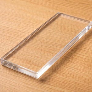 May include: A rectangular, clear acrylic block with smooth, polished edges and a flat surface. The object is transparent, allowing light to pass through its solid, thick structure, presenting a clean and simple geometric form for display purposes.