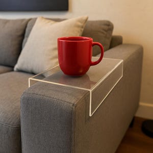 May include: A clear acrylic sofa arm tray holds a red mug on a gray upholstered sofa. The tray is rectangular and designed to fit over the arm of a couch. A beige pillow rests on the sofa behind the mug.