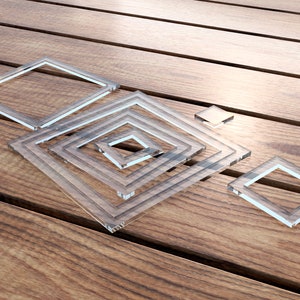 May include: A set of clear glass squares of varying sizes, arranged in a concentric pattern on a wooden surface.