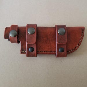 Op de afbeelding: Een bruine leren schede voor een mes met drie riemen en metalen drukknopen. De schede heeft een gestikte rand en een gebogen vorm.