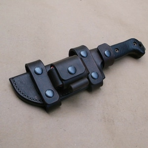 Könnte beinhalten: Eine schwarze Lederscheide mit einer braunen Lederschlaufe und Metalldruckknöpfen. Die Scheide ist für ein großes Messer mit schwarzem Griff konzipiert.