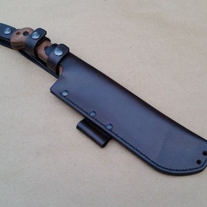 Könnte beinhalten: Eine braune Lederscheide mit einem Gürtelschlaufe und einem Druckknopfverschluss. Die Scheide ist für ein großes Messer oder eine Machete ausgelegt.
