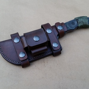 Peut inclure: Un fourreau en cuir marron avec un couteau noir à l'intérieur. Le fourreau a un rabat avec une fermeture à pression et plusieurs rivets métalliques. Le couteau a un manche noir enveloppé de paracorde verte.