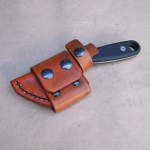 Peut inclure: Un étui à couteau en cuir marron avec un couteau noir à l'intérieur. L'étui comporte trois boutons-pression noirs et une couture cousue.