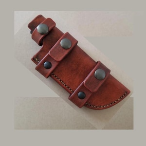 May include: A brown leather knife sheath with three straps and metal snaps. The sheath has a stitched edge and a curved shape.