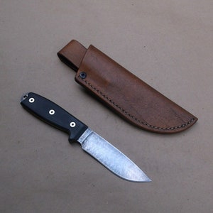 K&ouml;nnte beinhalten: Eine braune Ledertasche f&uuml;r ein Messer mit einem gen&auml;hten Rand und ein schwarzes Messer mit einem schwarzen Griff und einer silbernen Klinge.