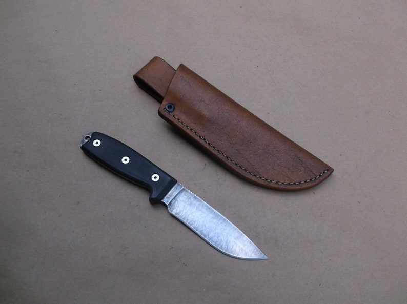 K&ouml;nnte beinhalten: Eine braune Ledertasche f&uuml;r ein Messer mit einem gen&auml;hten Rand und ein schwarzes Messer mit einem schwarzen Griff und einer silbernen Klinge.