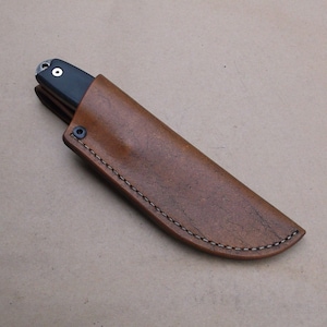 K&ouml;nnte beinhalten: Eine braune Ledertasche f&uuml;r ein Messer mit einem gen&auml;hten Rand und einem Druckknopfverschluss.