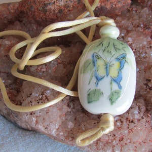 May include: A white ceramic pendant featuring a hand-painted butterfly design in shades of blue, yellow, and green. The pendant is attached to a yellow cord.