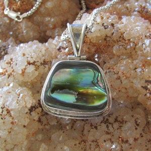 May include: A silver pendant with a green, blue, and gold abalone shell inlay. The pendant is on a silver chain and is resting on a bed of white crystals.