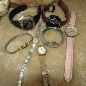 May include: A collection of seven wristwatches with different styles and bands. The watches have silver, gold, and black bands, and some have white faces with black numbers. One watch has a black band and a black face with white numbers.