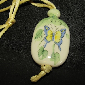May include: A white ceramic pendant with a yellow and blue butterfly painted on it. The pendant is attached to a yellow cord.