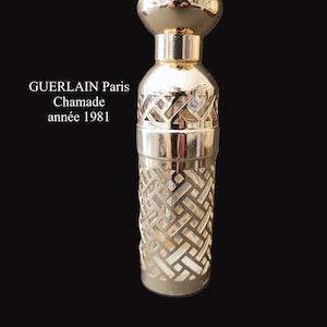 Könnte beinhalten: Eine Vintage-Parfümflasche mit goldfarbenem, geometrisch gemustertem Äußeren. Die Flasche hat einen abgerundeten Deckel und ein dekoratives, ausgeschnittenes Design. Der Text "GUERLAIN Paris Chamade année 1981" ist auf der Flasche aufgedruckt.