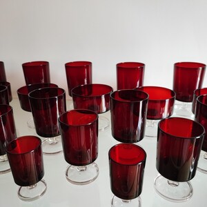 Könnte beinhalten: Eine Sammlung rubinroter Gläser, darunter Stielgläser und Becher. Die Gläser haben eine tiefe, satte Farbe und sind auf einer reflektierenden Oberfläche angeordnet. Die Vielfalt der Formen deutet auf ein Getränkeservice hin.