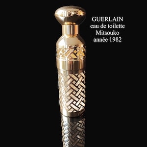 Op de afbeelding: Een goudkleurige parfumfles met een geometrisch patroon en een afgeronde bovenkant. De fles heeft de tekst "GUERLAIN eau de toilette Mitsouko année 1982". De fles wordt weerspiegeld op een zwart oppervlak.