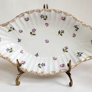 May include: White porcelain serving dish with a scalloped edge and gold trim, decorated with small pink and purple floral designs. The dish is displayed on a decorative brass stand, showcasing its elegant design. A charming piece for serving or display.