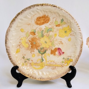 Puede incluir: Un plato decorativo vintage con un diseño floral. El plato presenta un fondo crema con flores amarillas, naranjas y rosas. El borde tiene un ribete dorado. El plato se exhibe sobre un soporte negro.