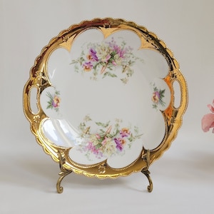 May include: A white porcelain serving dish with a gold rim and floral design. The dish has two handles and is on a gold metal stand.