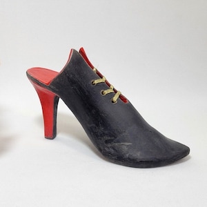 Könnte beinhalten: Schwarzer und roter High-Heel-Schuh-Skulptur mit goldfarbenen Schnürsenkeln. Der Schuh hat eine spitze Zehenpartie und einen roten Absatz. Das Innere des Schuhs ist rot. Im Hintergrund befindet sich ein dekoratives Blumenarrangement.