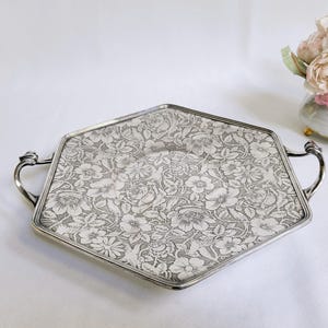 Może przedstawiać: Srebrzysta, sześciokątna taca do serwowania z kwiatowymi detalami. Taca ma dwa zakrzywione uchwyty i szczegółowy kwiatowy wzór na powierzchni. Mały wazon z kwiatami znajduje się w tle.