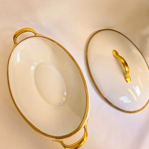 May include: An oval white porcelain serving dish with two gold-toned handles and a matching oval lid featuring a gold-toned handle. The rim of the dish and the edge of the lid are decorated with a thin, intricate gold-colored patterned border.