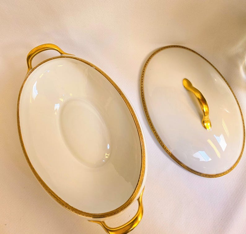 May include: An oval white porcelain serving dish with two gold-toned handles and a matching oval lid featuring a gold-toned handle. The rim of the dish and the edge of the lid are decorated with a thin, intricate gold-colored patterned border.
