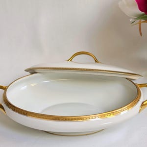 May include: A white porcelain serving dish with a matching lid, featuring two gold-toned handles on the sides and a gold-toned handle on the lid. The rim of the dish and the edge of the lid are decorated with a detailed gold geometric pattern.
