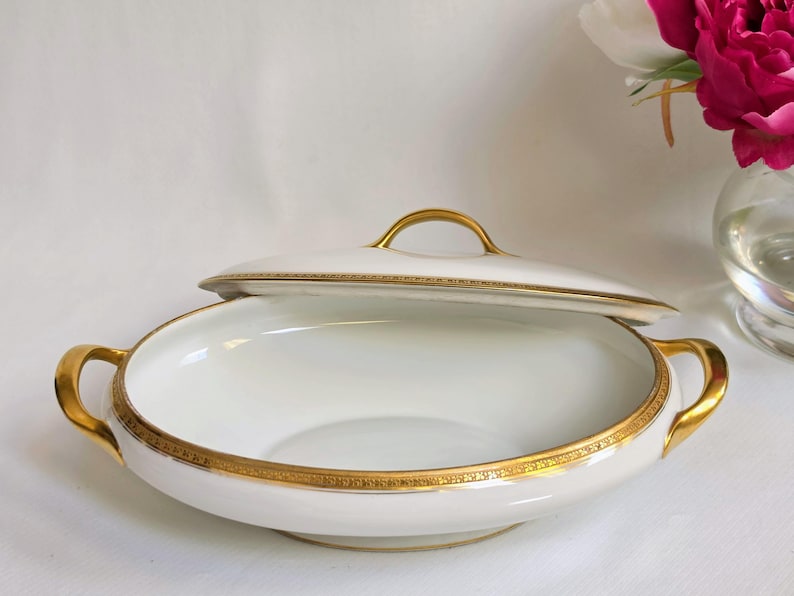 May include: A white porcelain serving dish with a matching lid, featuring two gold-toned handles on the sides and a gold-toned handle on the lid. The rim of the dish and the edge of the lid are decorated with a detailed gold geometric pattern.
