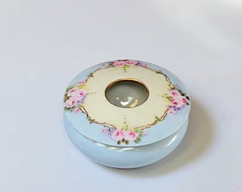 May include: A round, light blue ceramic trinket box with a floral design. The lid features a cream-colored center with a gold rimmed opening and a border of pink roses and green leaves. The box is a decorative item.