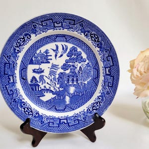 May include: A blue and white ceramic plate with a Willow pattern design, displayed on a dark wooden stand. The plate features a detailed landscape scene with pagodas, bridges, and birds. The plate is approximately 10 inches in diameter.