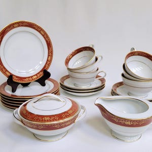 Puede incluir: Un juego de tazas de té y platillos rojos y dorados con una jarra de crema y un azucarero a juego. El juego está hecho de porcelana fina y presenta un delicado diseño floral.