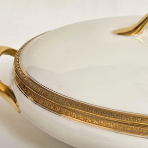 May include: An oval white porcelain serving dish with a lid, featuring a polished gold handle and a decorative gold band around the rim with an intricate repeating pattern. The dish has two curved gold side handles for lifting and carrying.