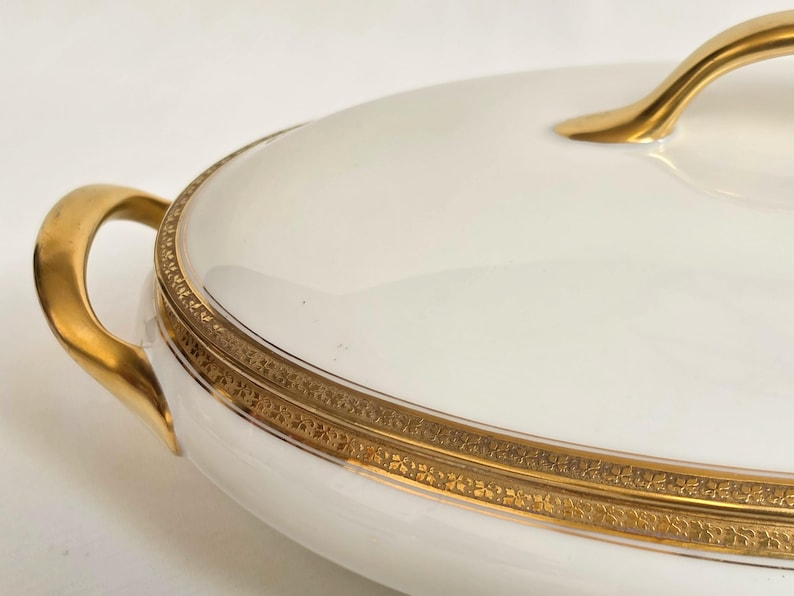 May include: An oval white porcelain serving dish with a lid, featuring a polished gold handle and a decorative gold band around the rim with an intricate repeating pattern. The dish has two curved gold side handles for lifting and carrying.