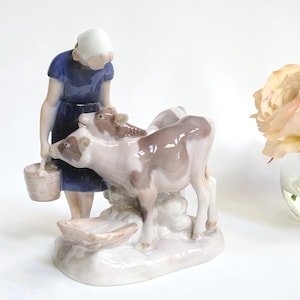 Op de afbeelding: Een porseleinen beeldje dat een vrouw in een blauwe jurk en witte kap voorstelt, die melk uit een emmer giet voor twee kalveren. De kalveren zijn bruin en wit, en de basis is wit met een trog.