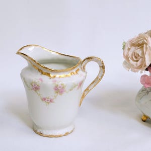 Op de afbeelding: Een witte porseleinen roomkannetje met een gouden rand en handvat, versierd met roze bloemmotieven. Het roomkannetje heeft een gebogen tuit en staat op een kleine voet. Een decoratieve vaas met roze en witte rozen staat op de achtergrond.