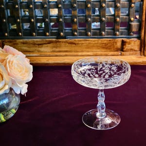 Puede incluir: Una copa de champán de cristal transparente grabado con un diseño floral. La copa tiene un cuenco ancho y poco profundo y un tallo retorcido. Un ramo de rosas melocotón en un cuenco de cristal está a la izquierda.