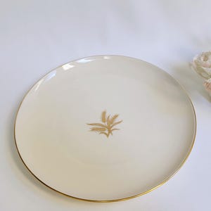 Peut inclure: Une assiette plate en céramique blanche avec un bord doré et un motif de blé doré au centre.