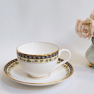 May include: White teacup and saucer set with a gold rim and a decorative band of navy blue, gold, and green floral design. The teacup has a curved handle. A vase of pink and cream roses is in the background.