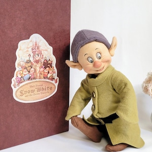 May include: A plush doll of a dwarf from the Disney film Snow White and the Seven Dwarfs. The dwarf is wearing a green coat with a black belt and a purple hat. The doll is sitting on a white surface. The doll is part of a collectible set.