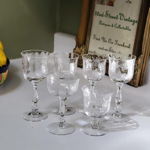 Puede incluir: Seis copas de cristal transparente grabadas con diseños florales. Las copas tienen un tallo delicado y una base redonda. Las copas están dispuestas sobre una superficie blanca, con un letrero enmarcado en el fondo. Plátanos y tomates en un cuenco decorativo.