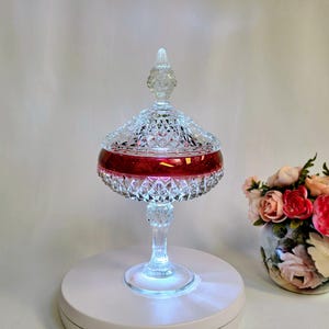 Puede incluir: Un cuenco de cristal transparente sobre pedestal con un patrón de corte de diamante y tapa a juego. El cuenco presenta una ancha banda horizontal de color rojo intenso. El objeto se apoya en una base circular e iluminado desde abajo.