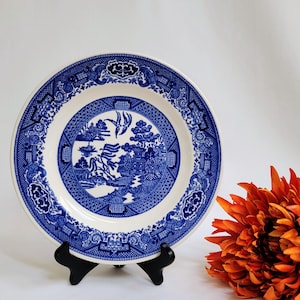 May include: A blue and white porcelain plate with a traditional willow pattern. The plate is on a black stand and has a white background with a blue border.