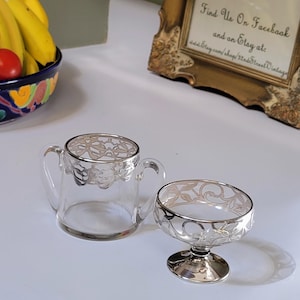 May include: A clear glass creamer and sugar bowl set with silver filigree trim. The creamer has two handles, and the sugar bowl sits on a pedestal base. The set is displayed on a white surface.