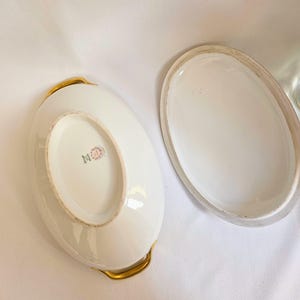 May include: An oval white porcelain serving dish with a matching lid, featuring gold-toned handles on the sides. The underside of the dish displays a maker's mark with the text Limoges France and a floral emblem in the center of the base.