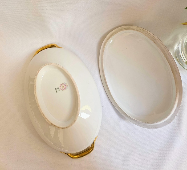 May include: An oval white porcelain serving dish with a matching lid, featuring gold-toned handles on the sides. The underside of the dish displays a maker's mark with the text Limoges France and a floral emblem in the center of the base.