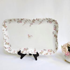 May include: A rectangular, white ceramic serving tray with a scalloped edge. The tray is decorated with pink and purple floral patterns. It is displayed on a black stand, with a small bouquet of roses in the background.