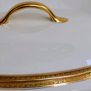 May include: A white porcelain serving dish lid with a smooth, glossy finish. The lid features a curved, polished gold handle at the top and a wide, decorative gold band around the lower edge with an intricate, repeating floral pattern.