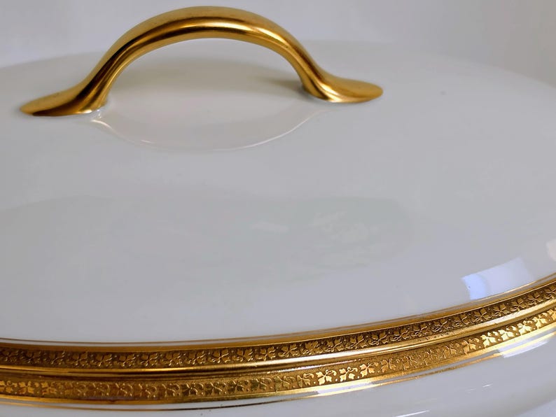 May include: A white porcelain serving dish lid with a smooth, glossy finish. The lid features a curved, polished gold handle at the top and a wide, decorative gold band around the lower edge with an intricate, repeating floral pattern.
