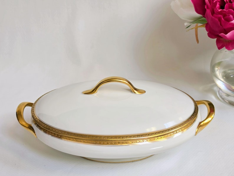 May include: An oval white porcelain serving dish with a matching lid, featuring a gold-toned handle on top and two gold-toned side handles. The rim of the lid and the base are decorated with a detailed, intricate gold band pattern.