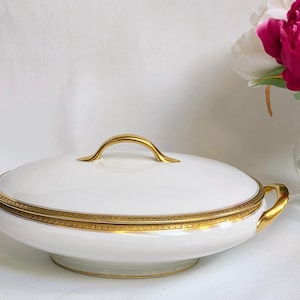 May include: A white porcelain serving dish with a lid, featuring a gold-toned handle and two gold-toned side handles. The rim of the lid and the base of the dish are decorated with a detailed gold band pattern. The dish has a smooth finish.